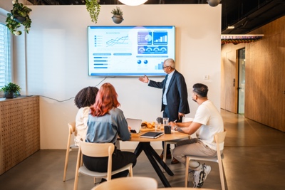Senior Multiracial Male Sales Executive Enthusiastically Presenting Financial Report to Diverse Colleagues in Modern Office